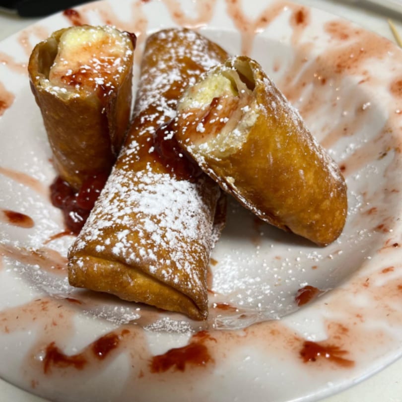 Three golden-brown dessert spring rolls, dusted with powdered sugar and drizzled with strawberry sauce.