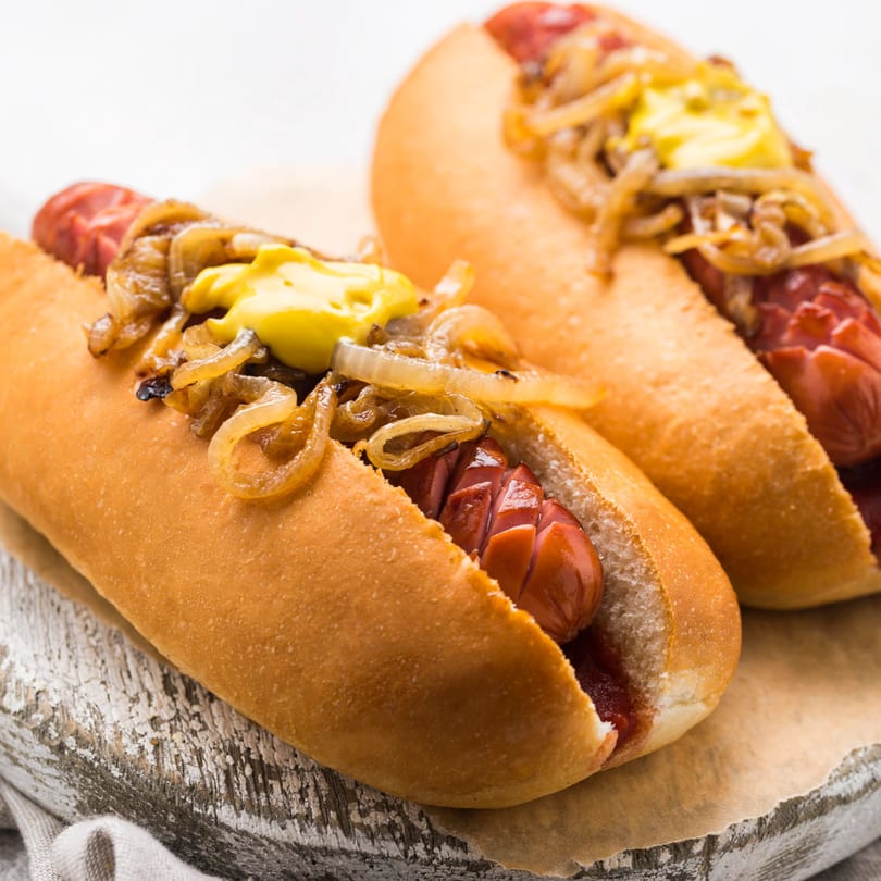 Two hot dogs with caramelized onions and mustard on buns, on a rustic wooden board.